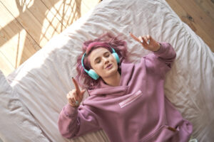 Adolescente com cabelo rosa usa fones de ouvido deitada na cama confortável ouvindo música.